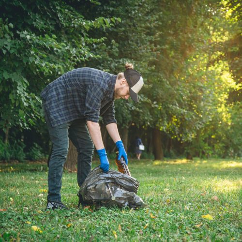A young man with a garbage bag in the forest cleans up plastic bottles, the concept of love for nature and care for the ecology, copy space.